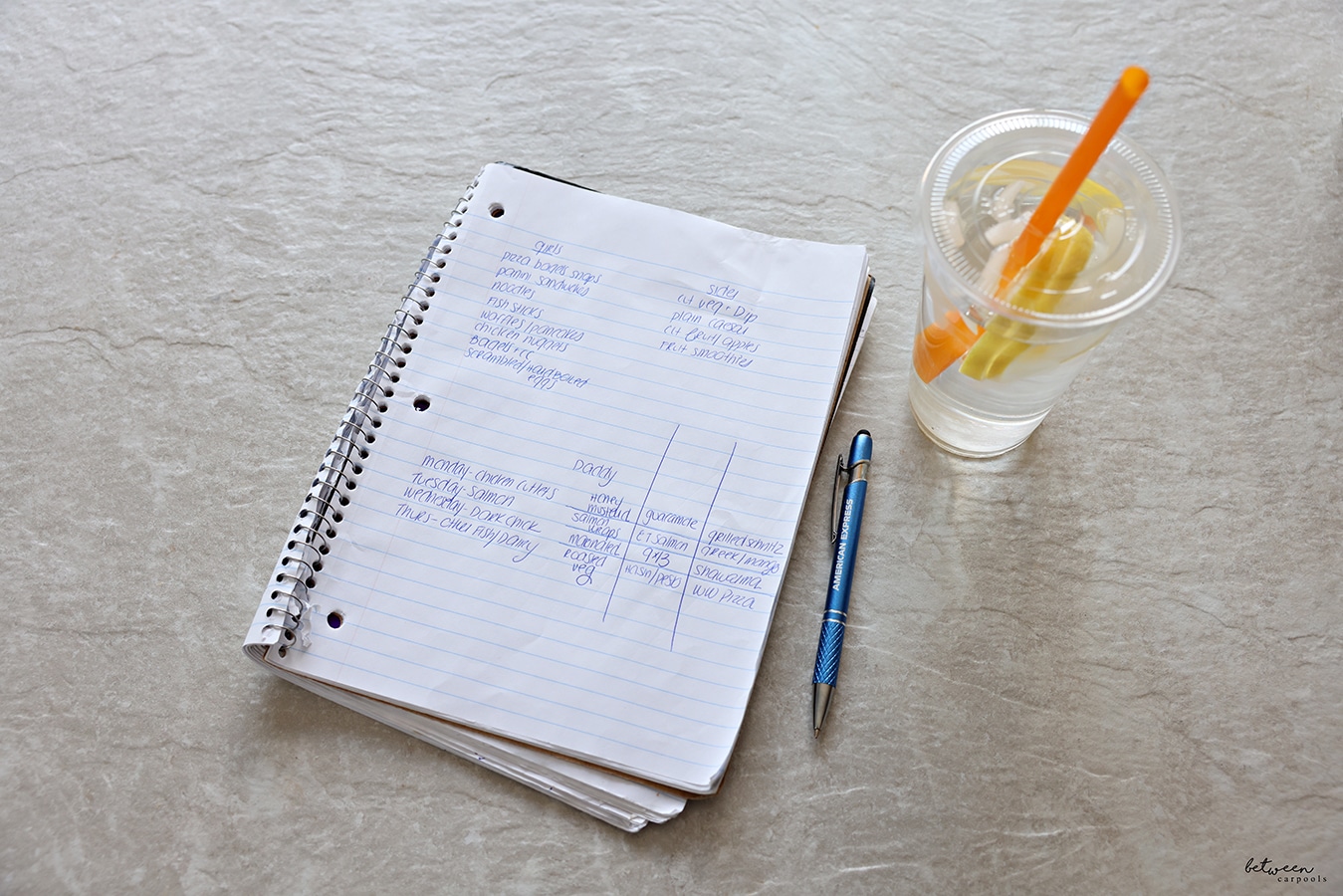 Handwritten menu plan in a spiral notebook. Pen and lemon water nearby