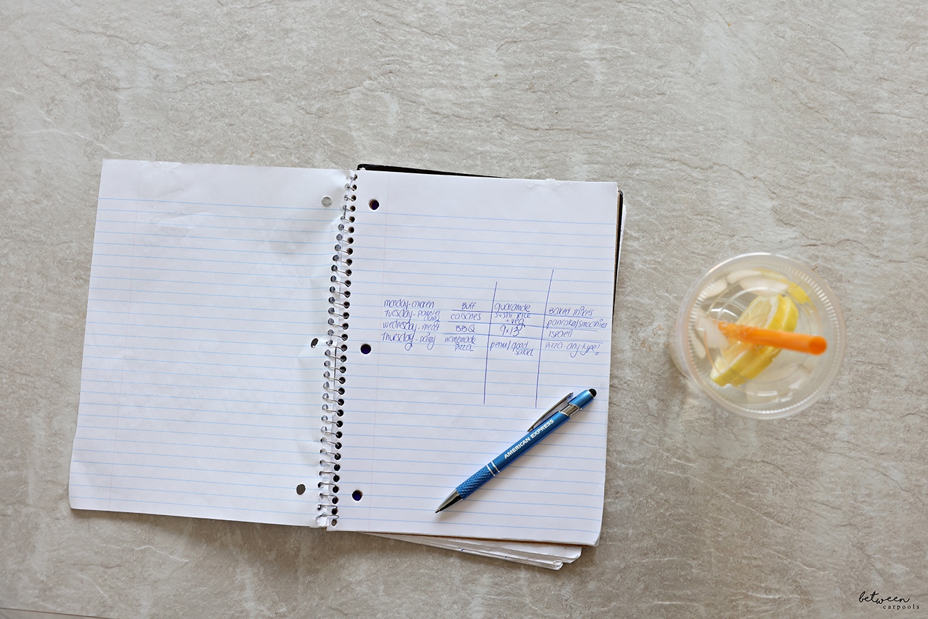 Handwritten menu plan in a spiral notebook. Pen and lemon water nearby
