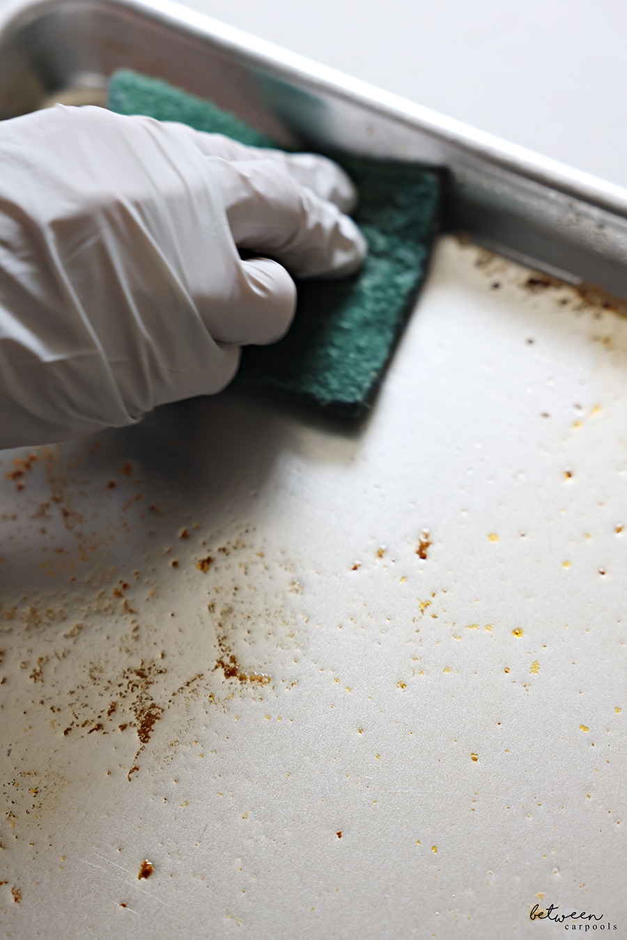Cleaning a metal sheet pan soaking with dish soap + water with a gloved hand and half a Scotch-Brite Heavy Duty Scour Pad