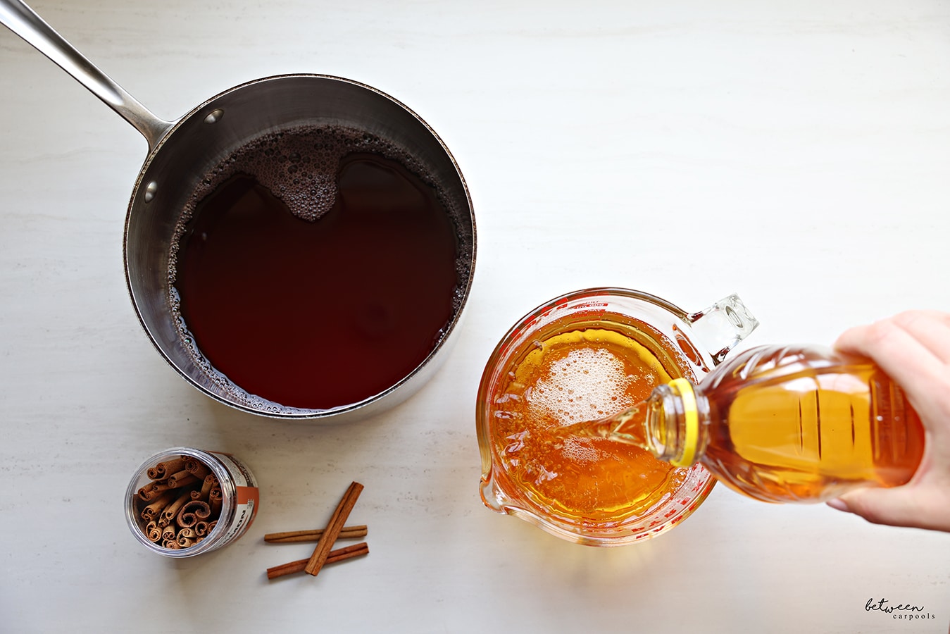 Cranberry juice in a pot, pouring apple juice into a Pyrex glass measuring cup, cinnamon sticks nearby