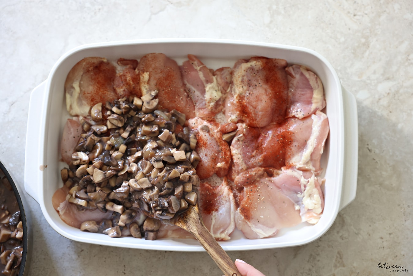 Adding sautéed mushrooms and onion over boneless chicken with salt, paprika, and pepper in a white oven to table baking dish.