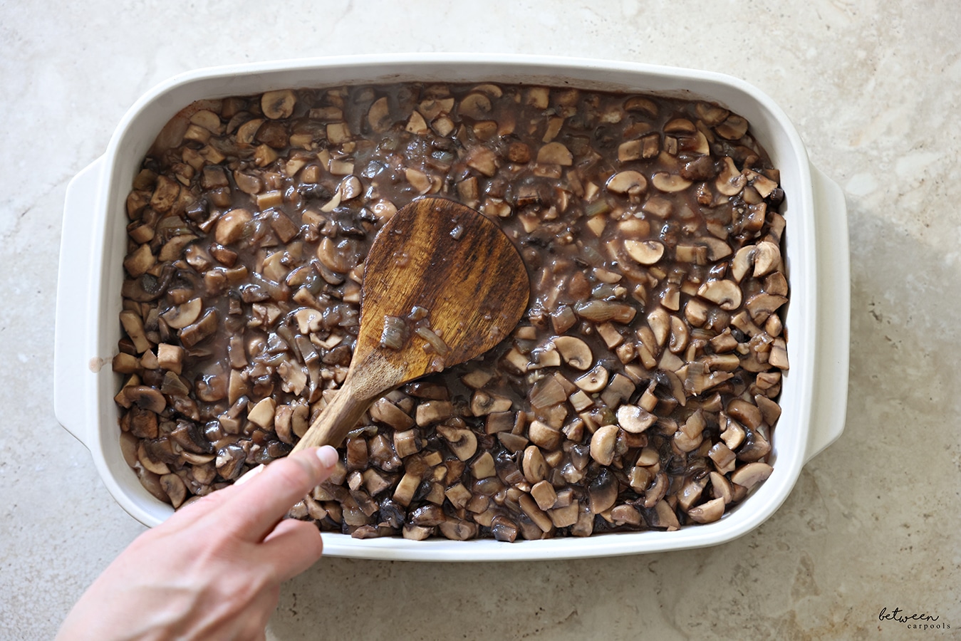 Spreading the sautéed mushrooms and onion over boneless chicken in a white oven to table baking dish with a wooden spoon.