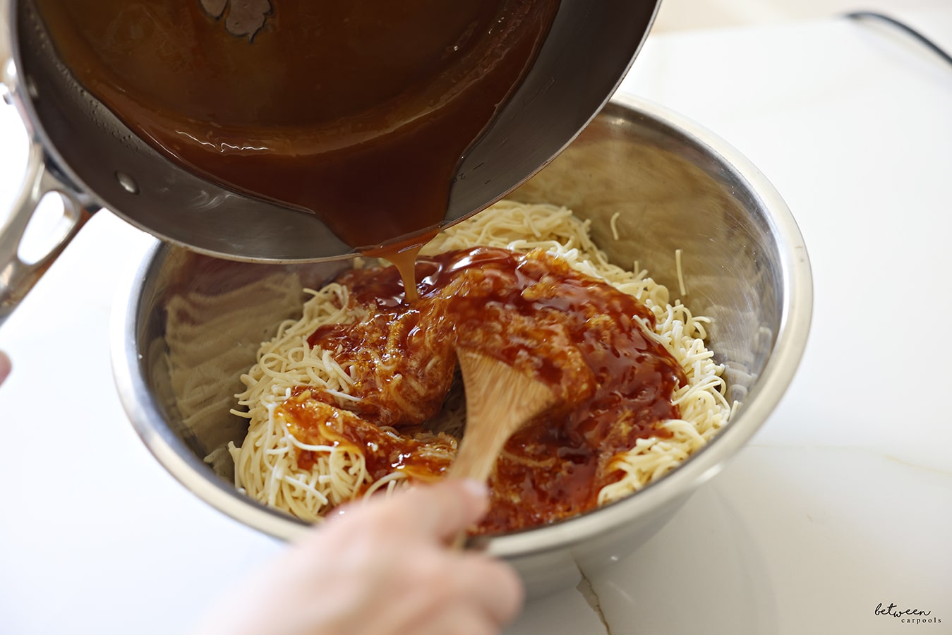 Pouring deeply golden oil and sugar into a metal bowl with cooked fine egg noodles.