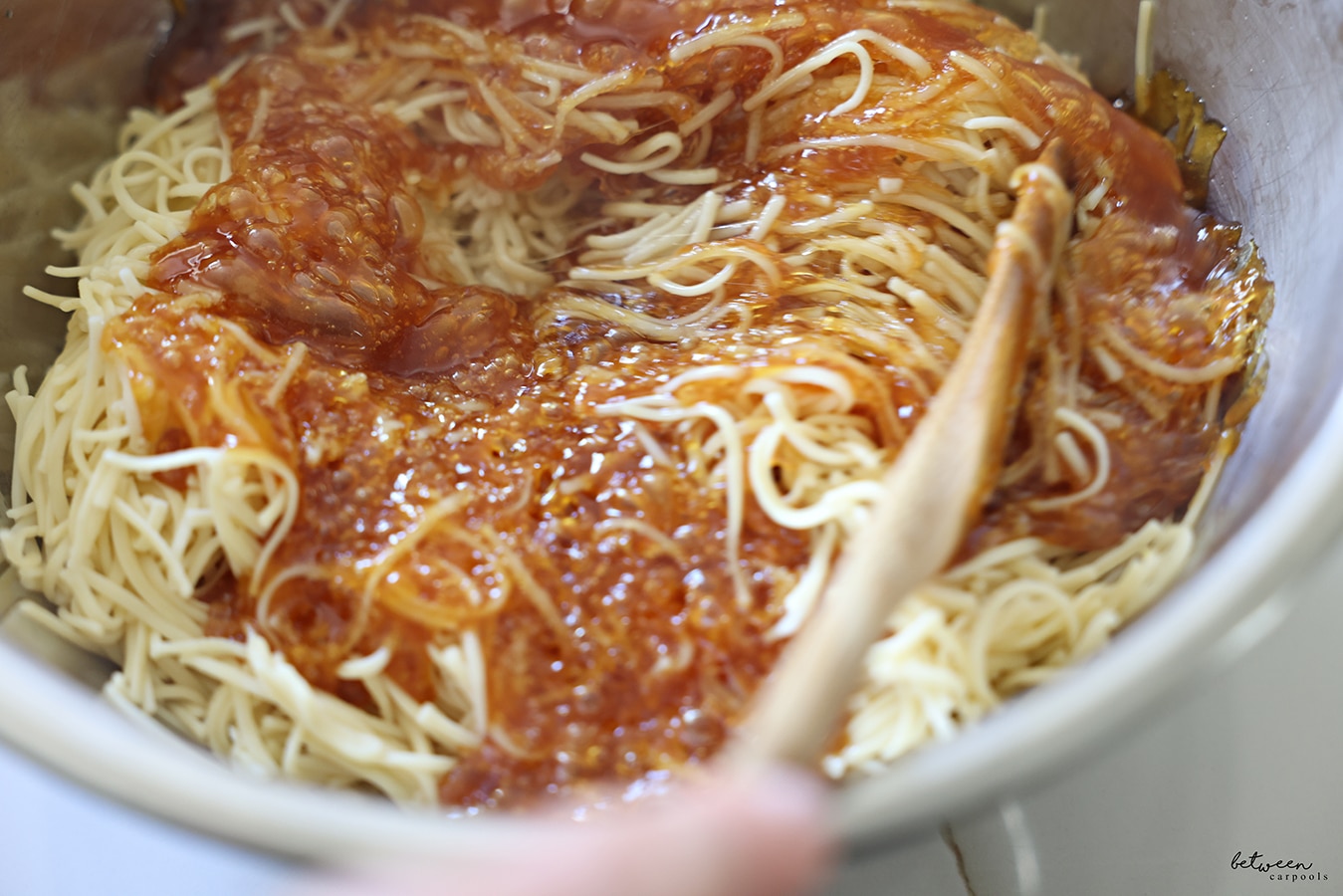 Mixing deeply golden oil and sugar with cooked fine egg noodles in a metal bowl with a wooden spoon.
