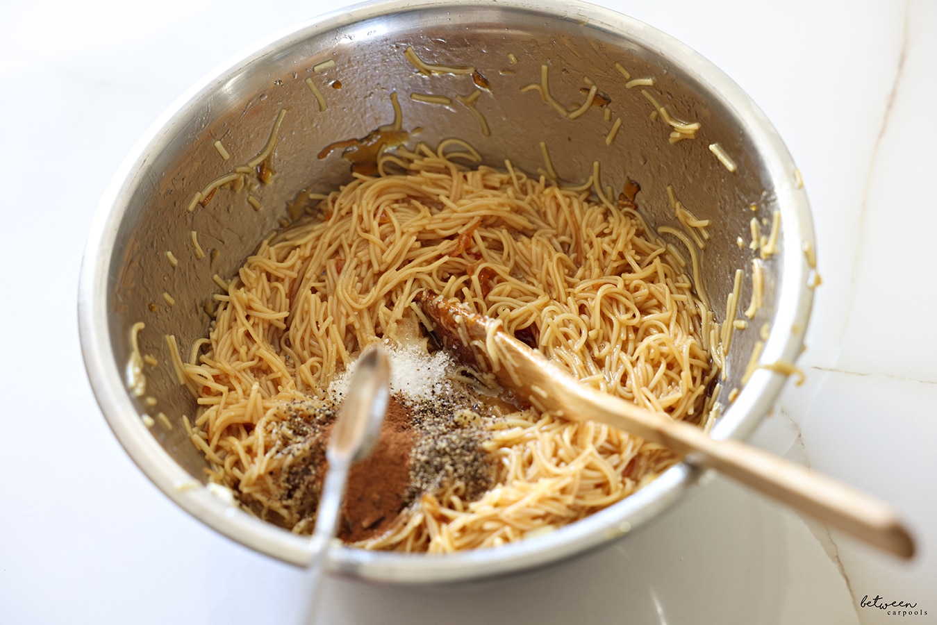 Adding spices to cooked fine egg noodles mixed with deeply golden oil and sugar in a metal bowl.