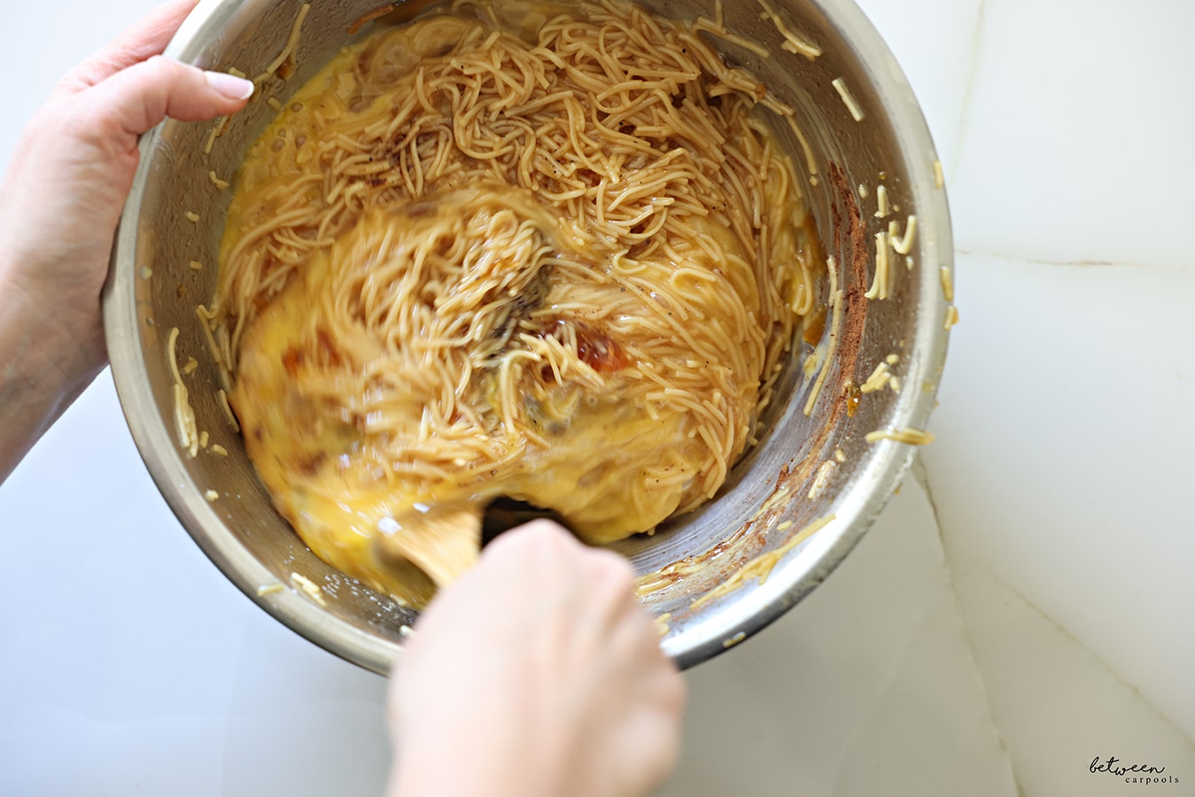 Stirring eggs with cooked fine egg noodles mixed with deeply golden oil and sugar plus spices in a metal bowl with a wooden spoon.