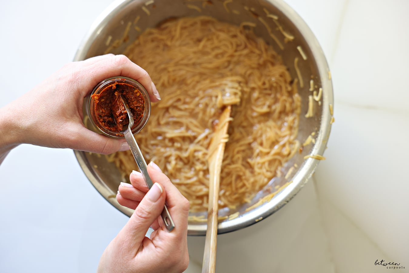 Adding a tbsp of red schug to cooked fine egg noodles mixed with deeply golden oil and sugar plus spices and eggs in a metal bowl with a wooden spoon.