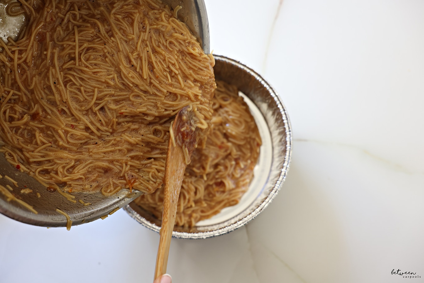 Pouring Yerushalmi Kugel mixture from a metal bowl into a round aluminum pan, with a wooden spoon.