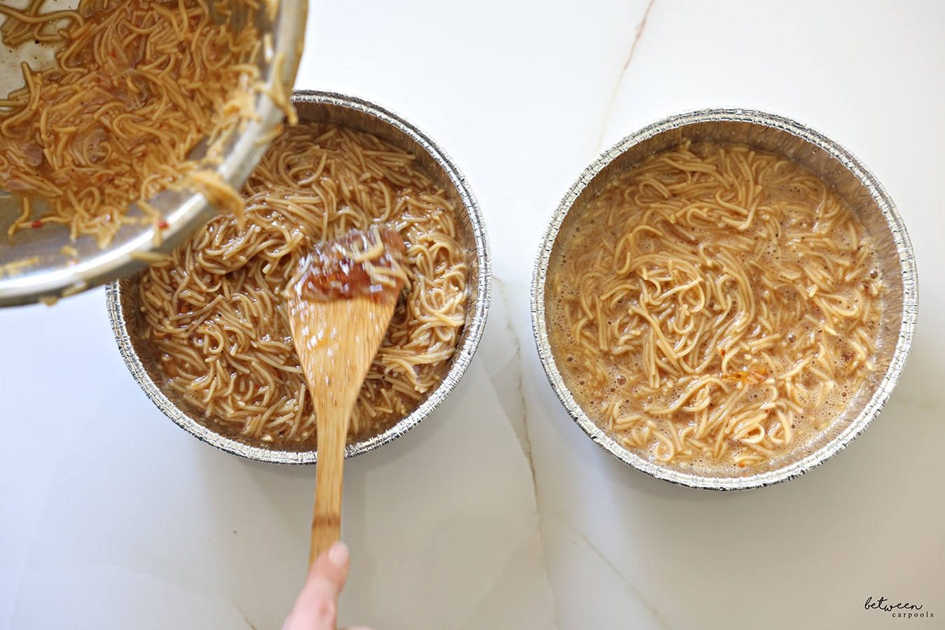 Pouring Yerushalmi Kugel mixture from a metal bowl into a second round aluminum pan, with a wooden spoon. Two round aluminum pans with Yerushalmi Kugel mixture on a kitchen counter.