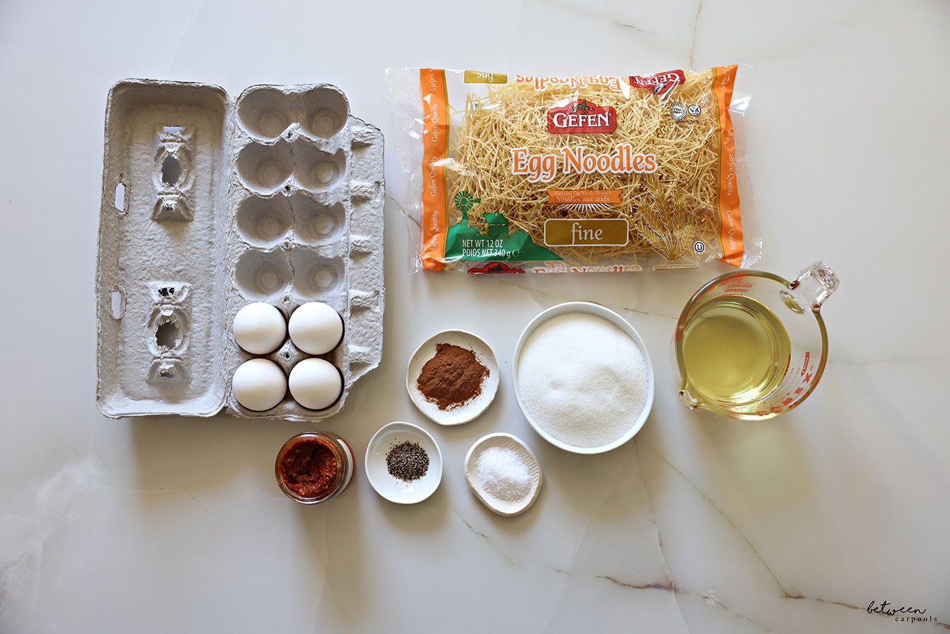 Ingredients for Yerushalmi Kugel. 4 eggs in a carton, 1 (12 oz) bag fine egg noodles, ¾ cup oil in a pyrex glass measuring cup, 1 ⅛ cups sugar in a bowl, salt, black pepper, cinnamon and a jar of red schug.
