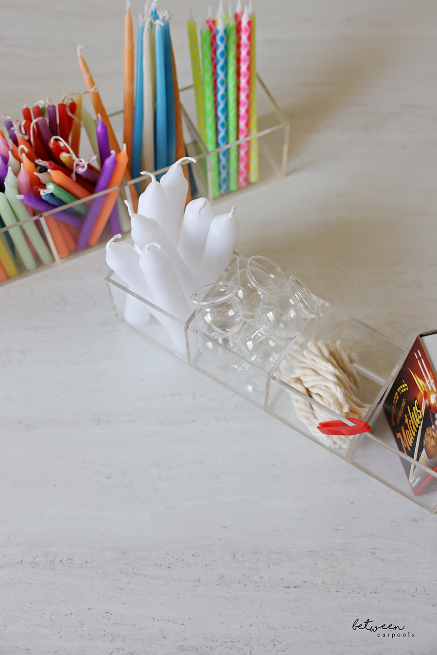 Closeup of 4-section acrylic organizers, filled. One with colored candles and one with Shabbos candles, Menorah oil glasses, wicks (plus a plastic tweezer) and a box of matches.