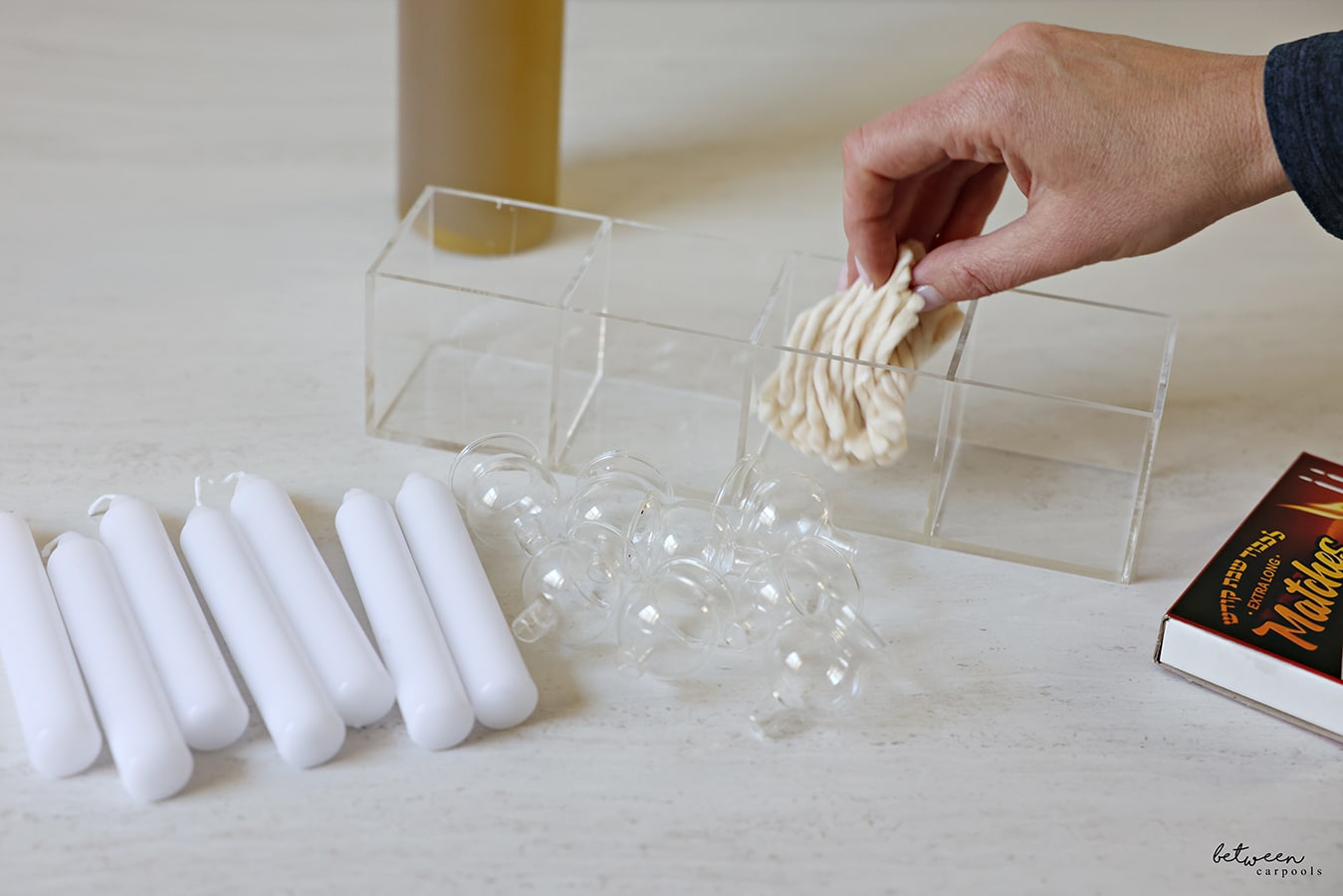Placing wicks into one section of a 4-section acrylic organizer. A condiment squeeze bottle with olive oil in the background. Shabbos candles, Menorah oil glasses and a box of matches.