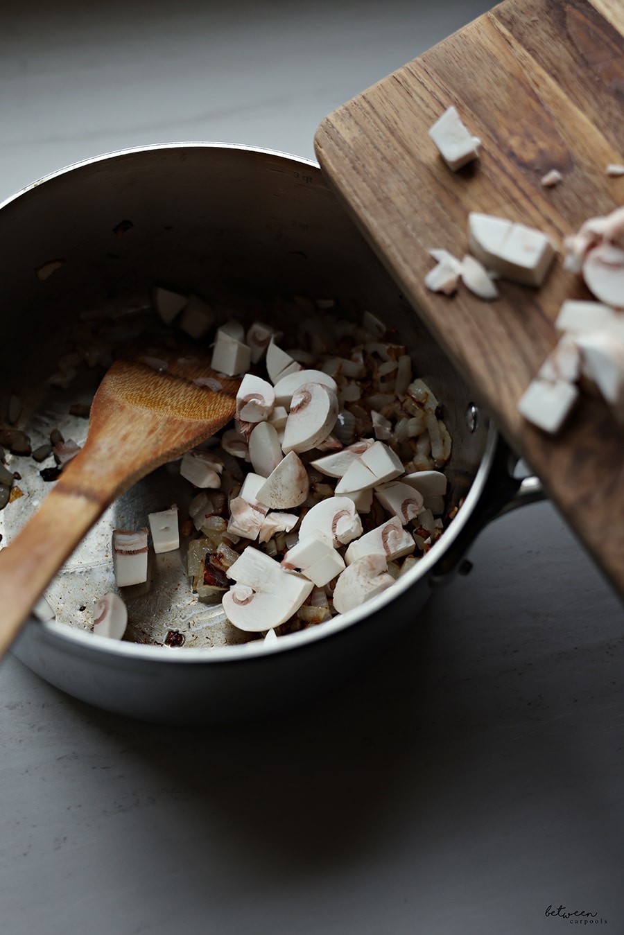 Adding sliced white onions to a saucepan with sautéed onions and a wooden spatula.