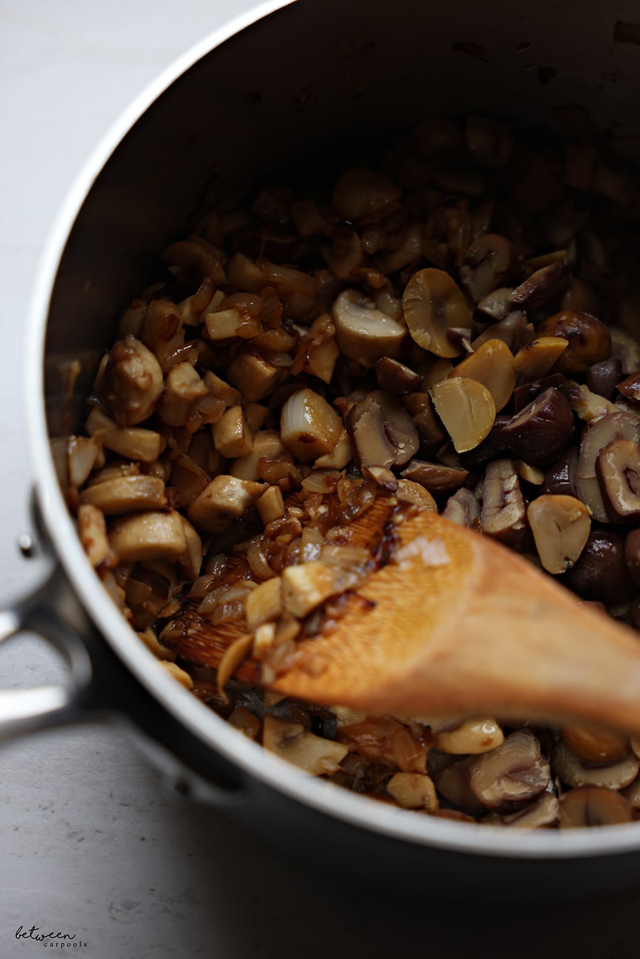 Closeup of sautéed onions, white mushrooms and garlic in a saucepan with a wooden spatula.
