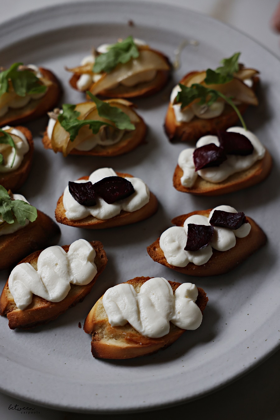 Bruschetta on a plate. Some with roasted pear slices, sunflower seeds, a drizzle of honey, and springs of arugula. Some topped with roasted beets.