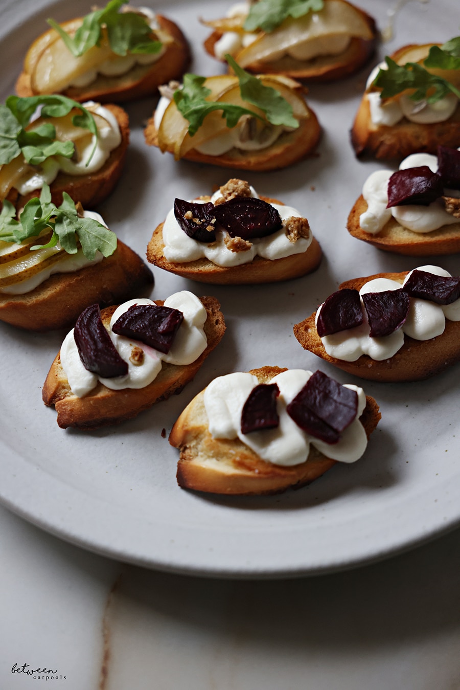 Bruschetta on a plate. Some with roasted pear slices, sunflower seeds, a drizzle of honey, and springs of arugula. Some topped with roasted beets and granola.