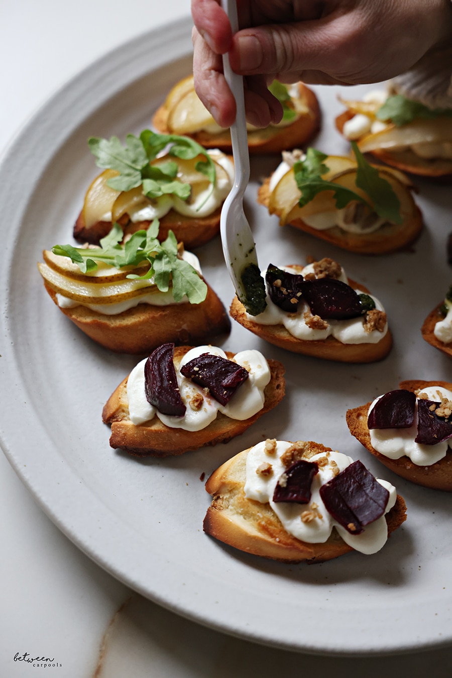 Bruschetta on a plate. Some with roasted pear slices, sunflower seeds, a drizzle of honey, and springs of arugula. Adding pesto to the bruschetta topped with roasted beets and granola.