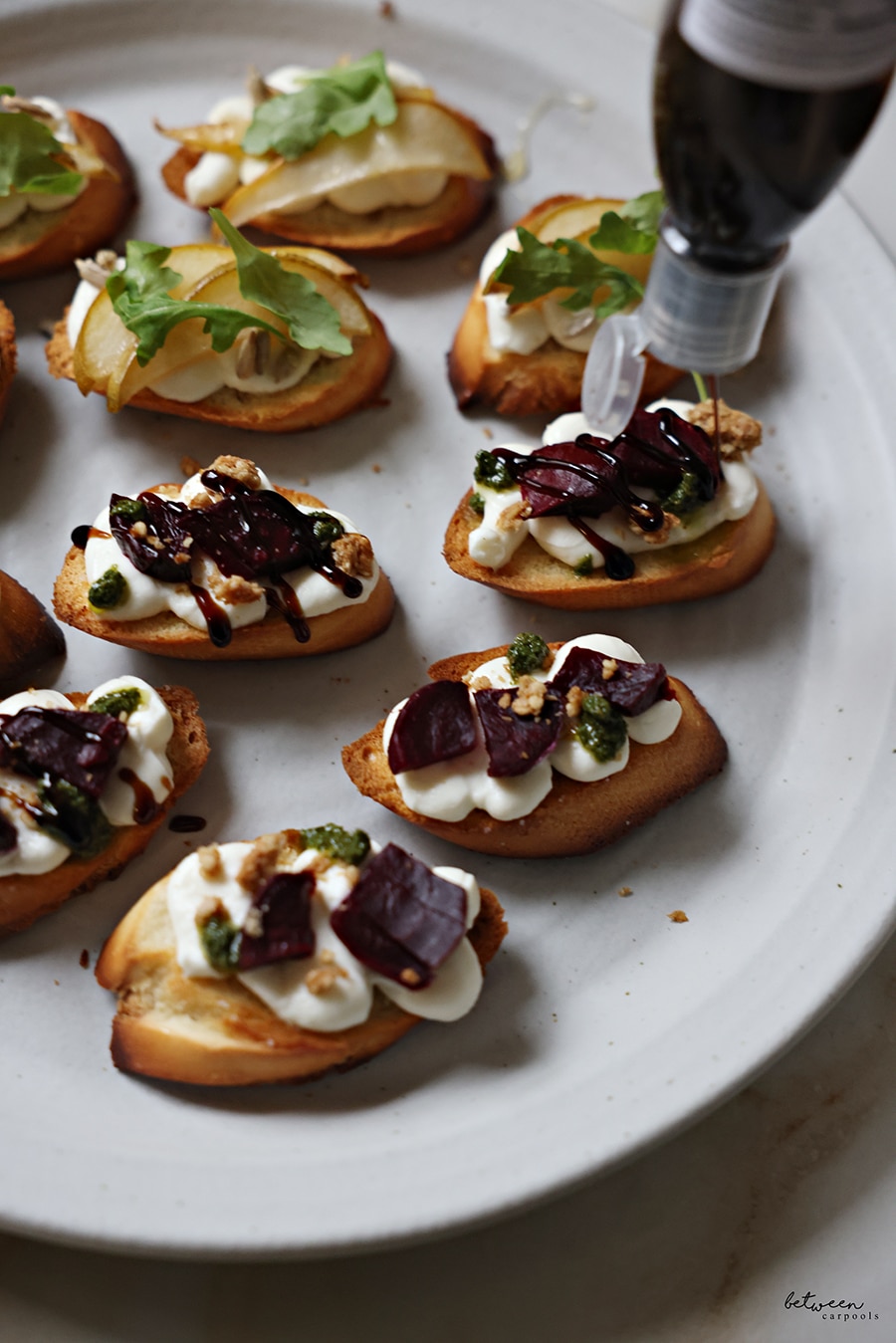 Bruschetta on a plate. Some with roasted pear slices, sunflower seeds, a drizzle of honey, and springs of arugula. Adding a drizzle of balsamic glaze to the bruschetta topped with roasted beets, granola, and pesto.