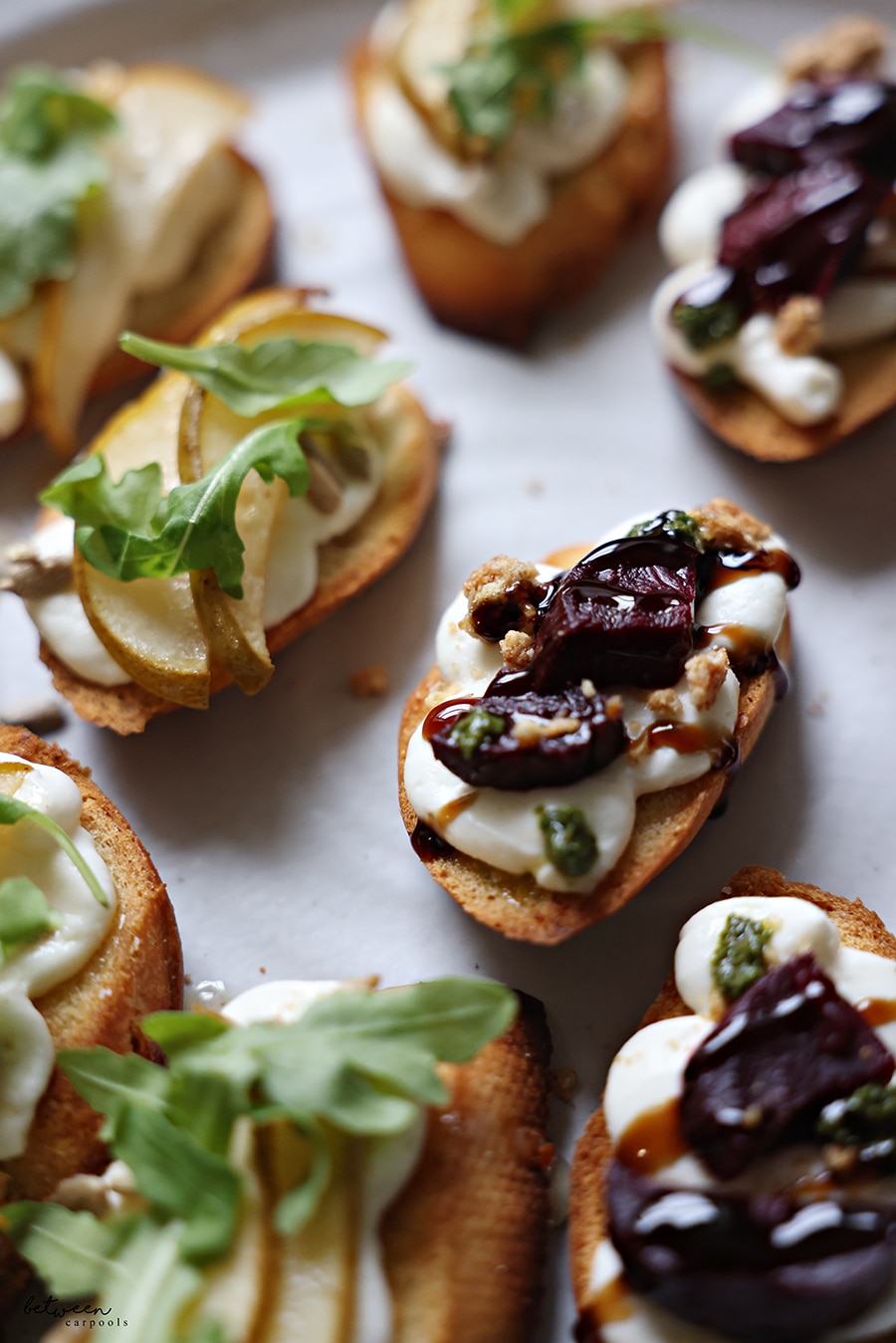 Closeup of bruschetta topped with roasted pear slices, sunflower seeds, a drizzle of honey, and springs of arugula - and bruschetta topped with roasted beets, granola, pesto, and a drizzle of balsamic glaze.
