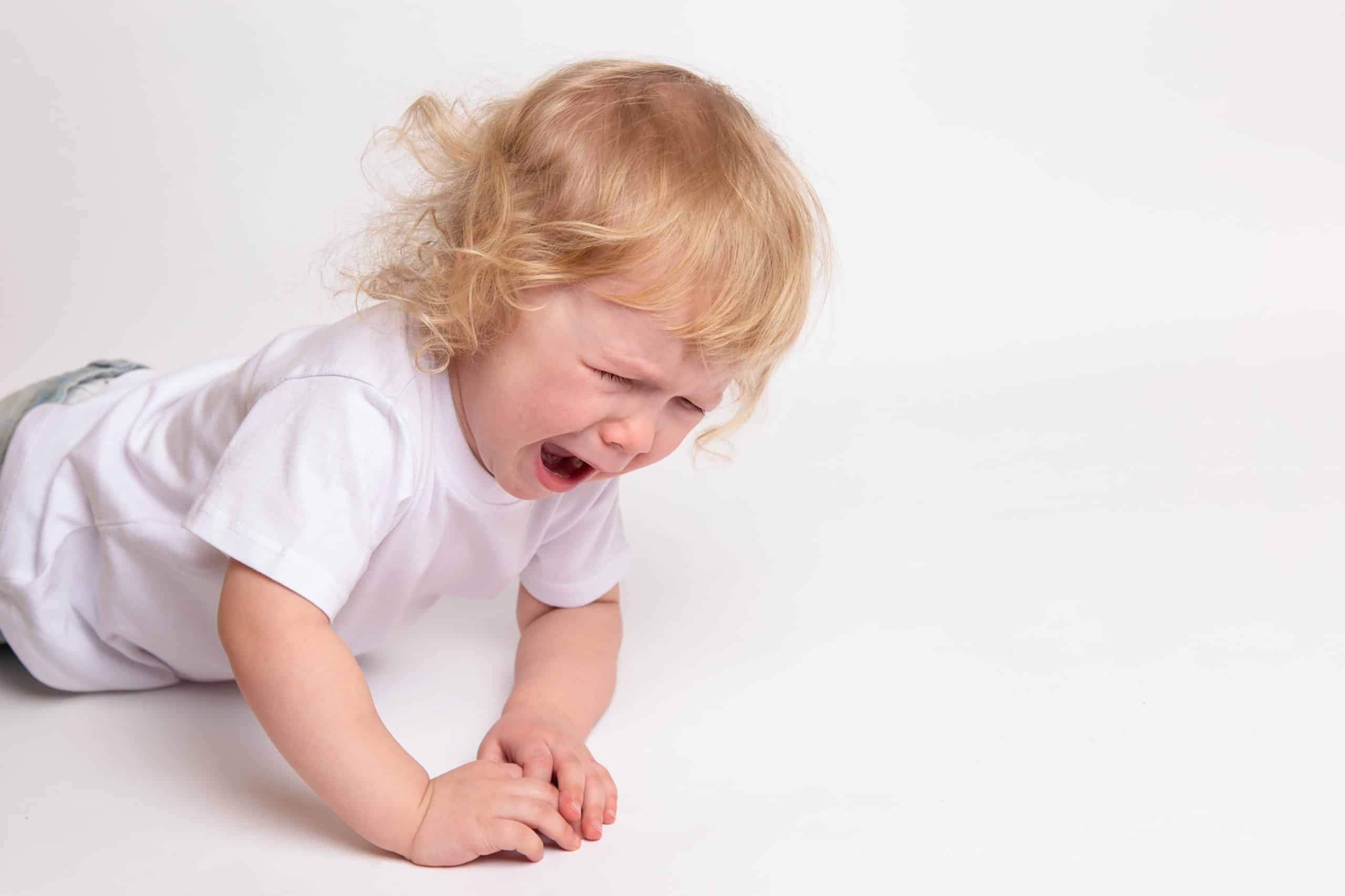A little curly crying child in a clean white T-shirt is lying on the floor and crying