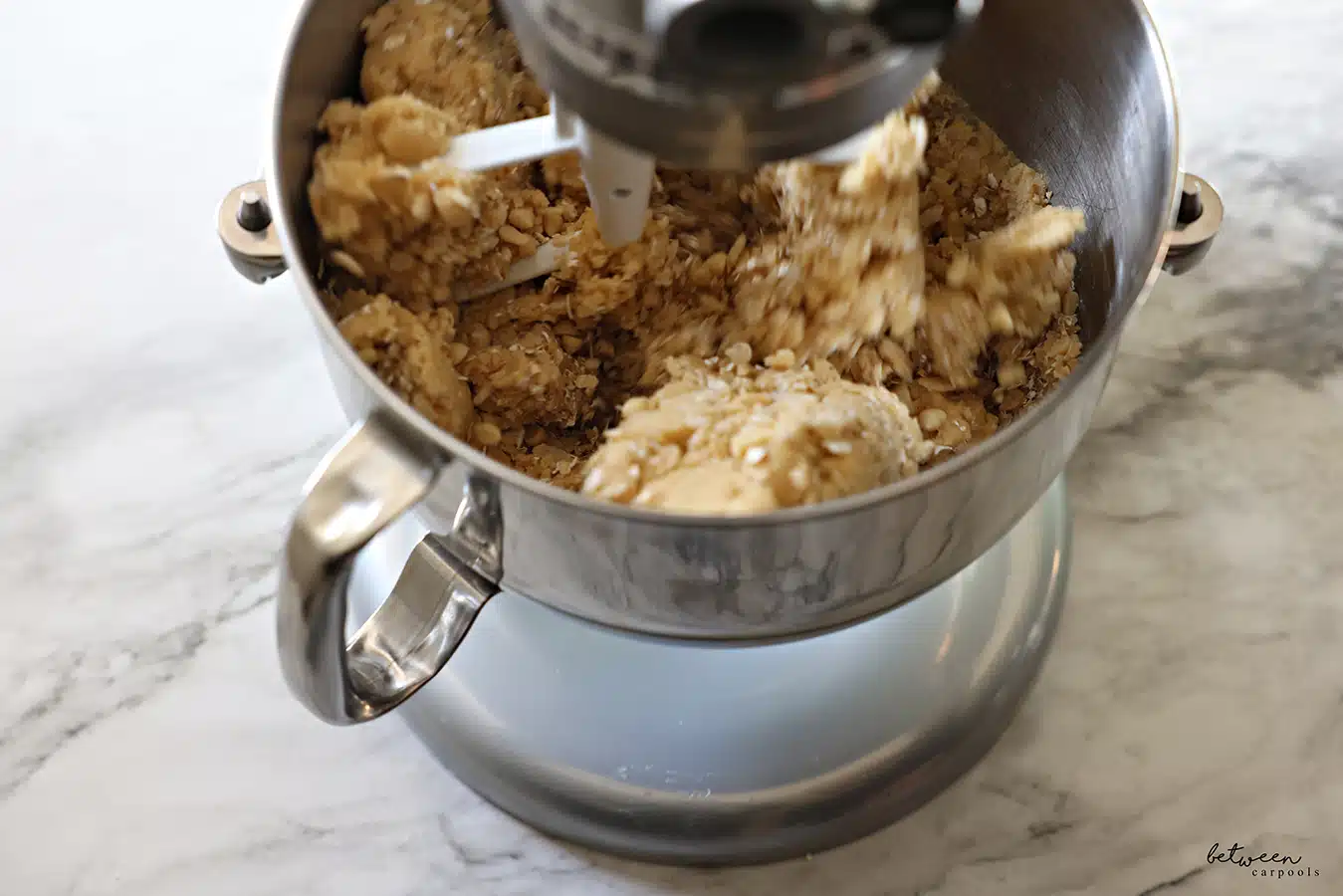 Mixing cookie dough with oats, Rice Krispies, and white chocolate chips, in the metal bowl of a Kitchen Aid mixer with the flat beater.