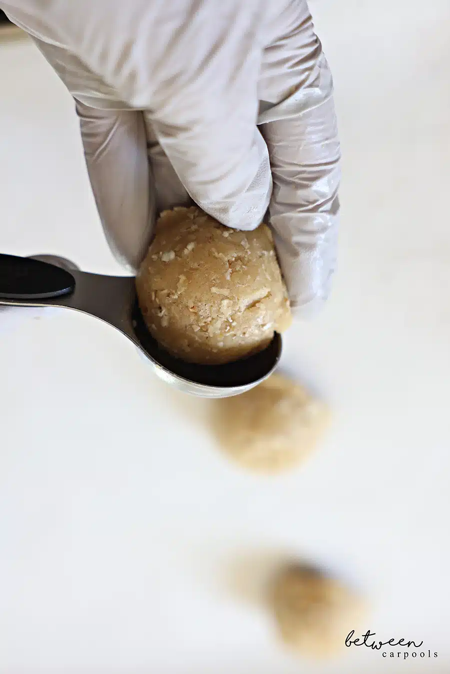Holding a ball of cookie dough with a gloved hand from a 1 tablespoon measuring spoon.
