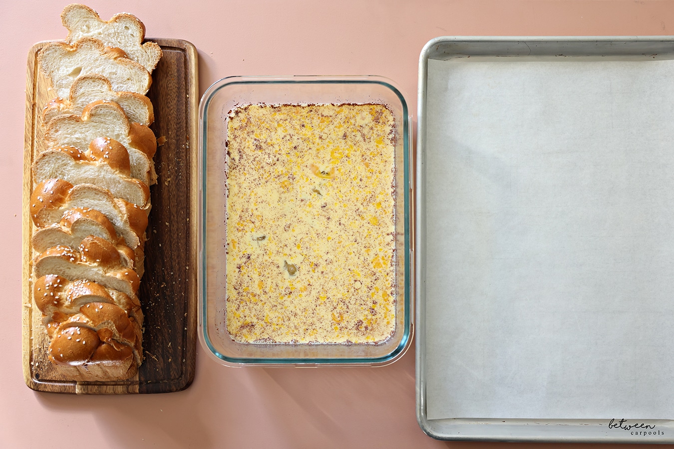 Sliced challah on a wooden board, French toast batter in a glass dish, and a parchment lined baking sheet.