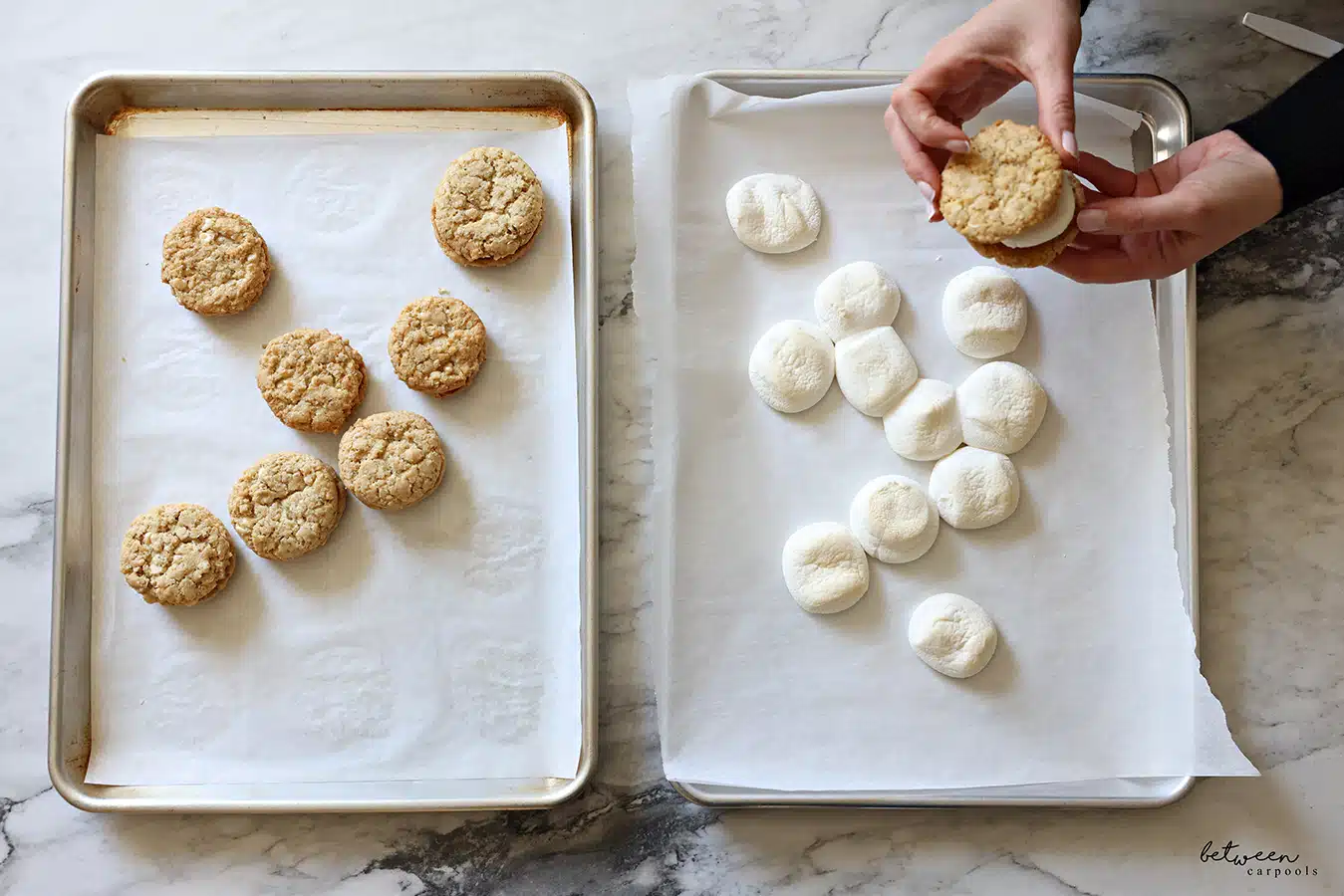 Pressing another cookie on top of a toasted marshmallow on a cookie. Two parchment lined baking sheets, one with cookies, one with toasted marshmallows.