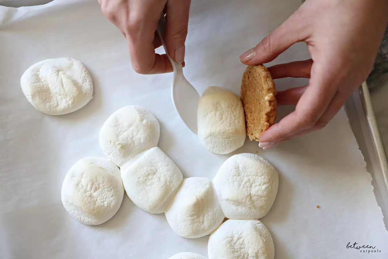 Scooping one toasted marshmallow - from a parchment lined cookie sheet of toasted marshmallows - against the flat side of one cookie.