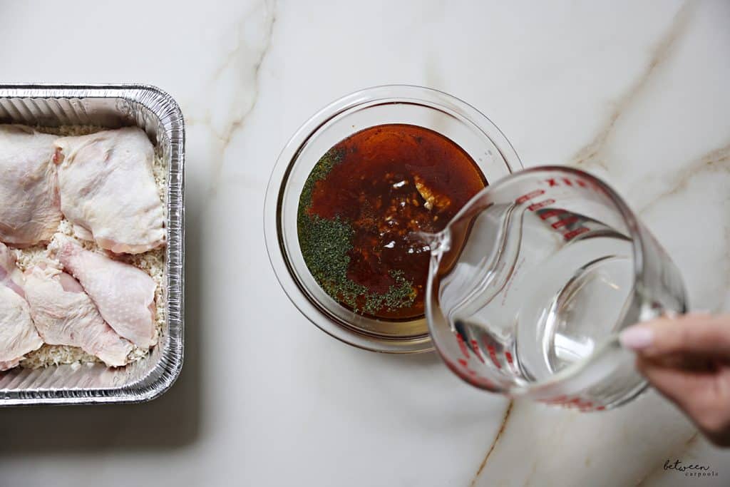 Adding boiling water to a glass bowl with oil, honey, soy sauce, garlic, orange juice, salt, pepper, paprika, and dried parsley. Chicken legs, cut in half, over rinsed raw rice in a 9x13 aluminum pan nearby.