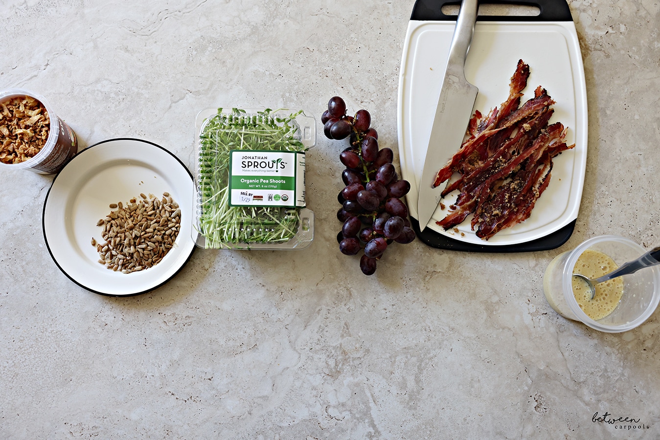 Pea Shoot Salad ingredients: container of crispy onions, sunflower seeds on a plate, a container of pea shoots, a bunch of grapes, Facon on a white plastic cutting board with a hast knife, salad dressing and a spoon in a container.