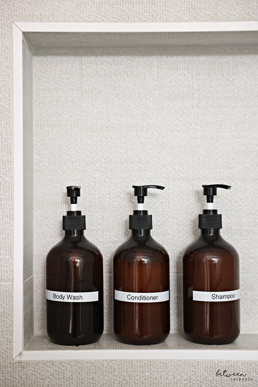 Three Amber Pump Bottles labeled Body Wash, Conditioner, and Shampoo, on a shower niche shelf.
