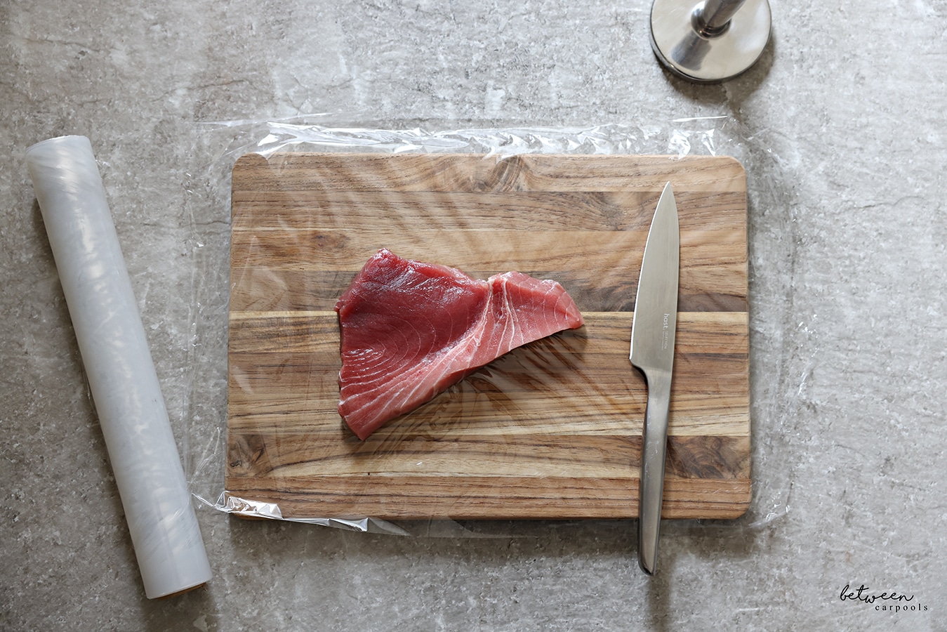 Fresh tuna and a hast knife on a wooden cutting board covered with plastic wrap. Roll of plastic wrap nearby.