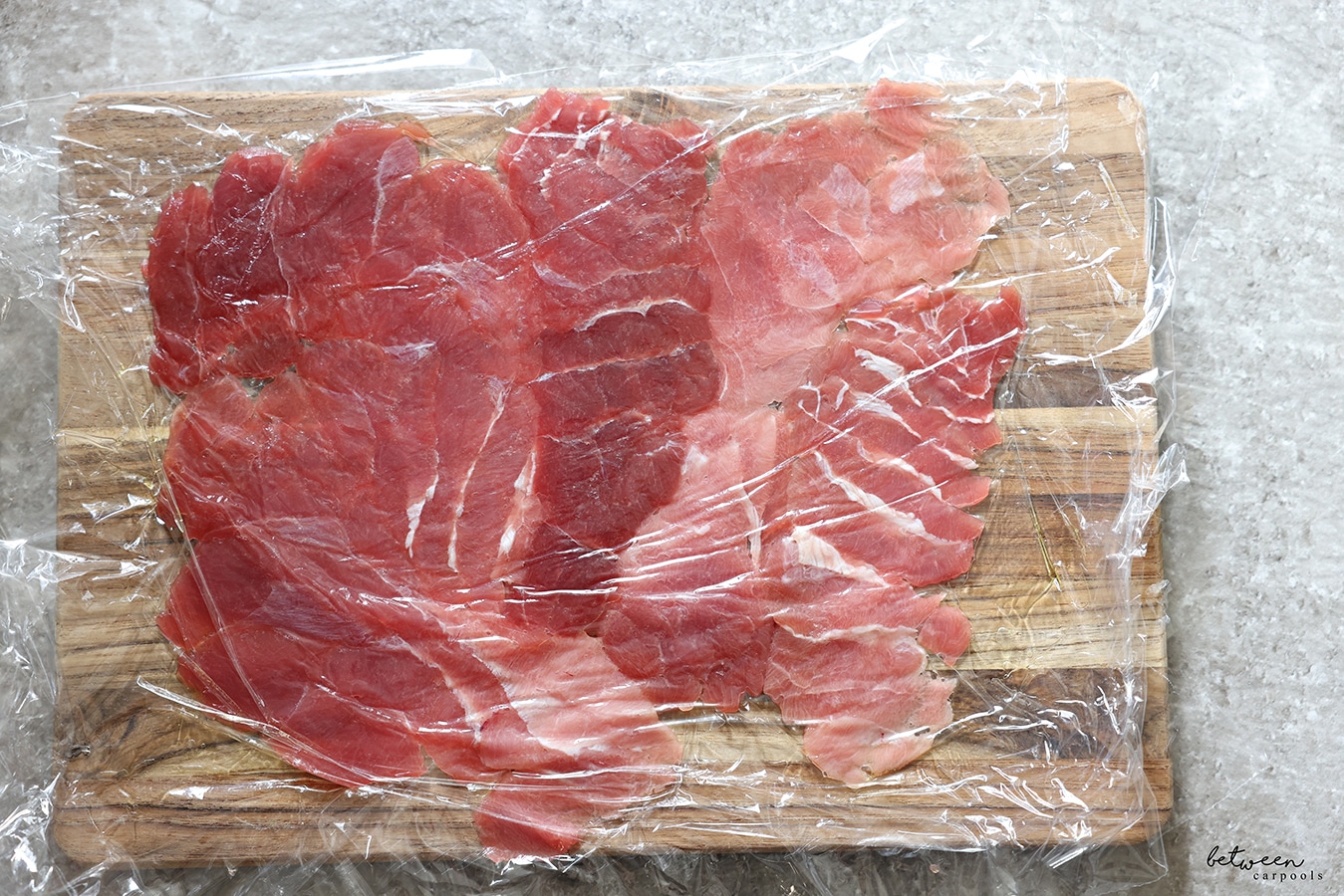 Pieces of fresh tuna pounded down between 2 layers of plastic wrap on a wooden cutting board.