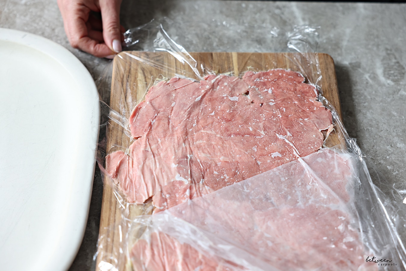 Removing one layer of plastic wrap from tuna pieces that have been pounded down between 2 layers of plastic wrap on a wooden cutting board and frozen.