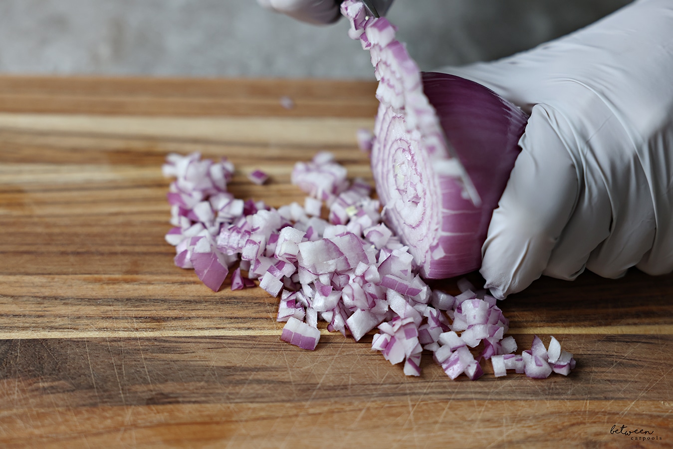 Dicing a red onion with gloved hands on a wooden cutting board.