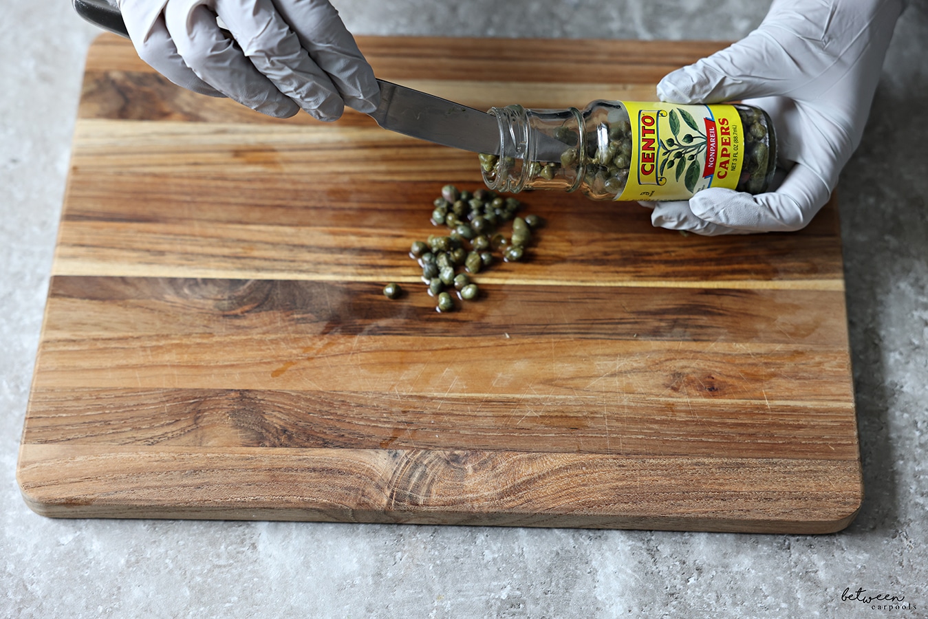 Pouring capers with gloved hands and a knife from its jar onto a wooden cutting board.