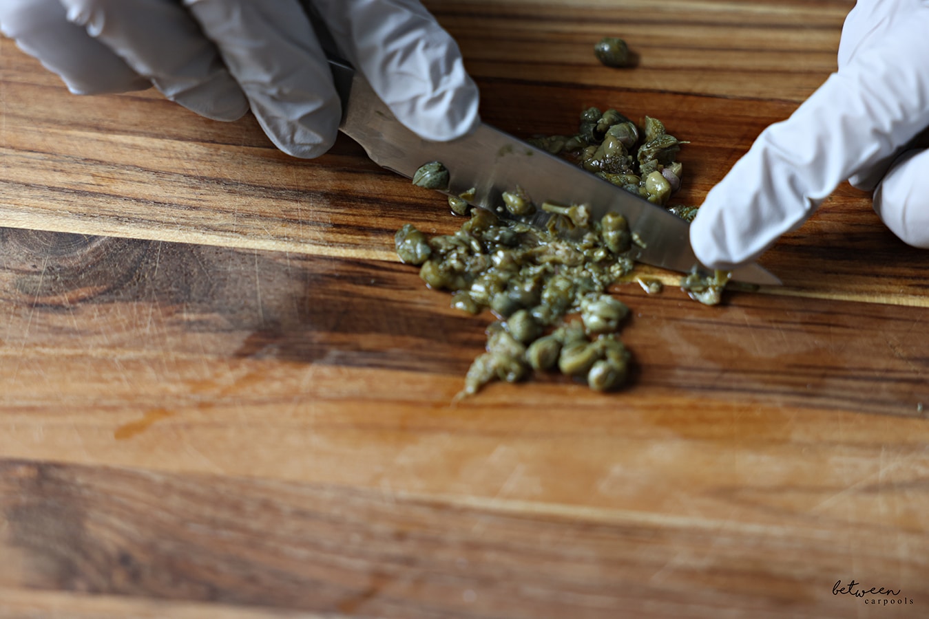 Dicing capers with gloved hands on a wooden cutting board.