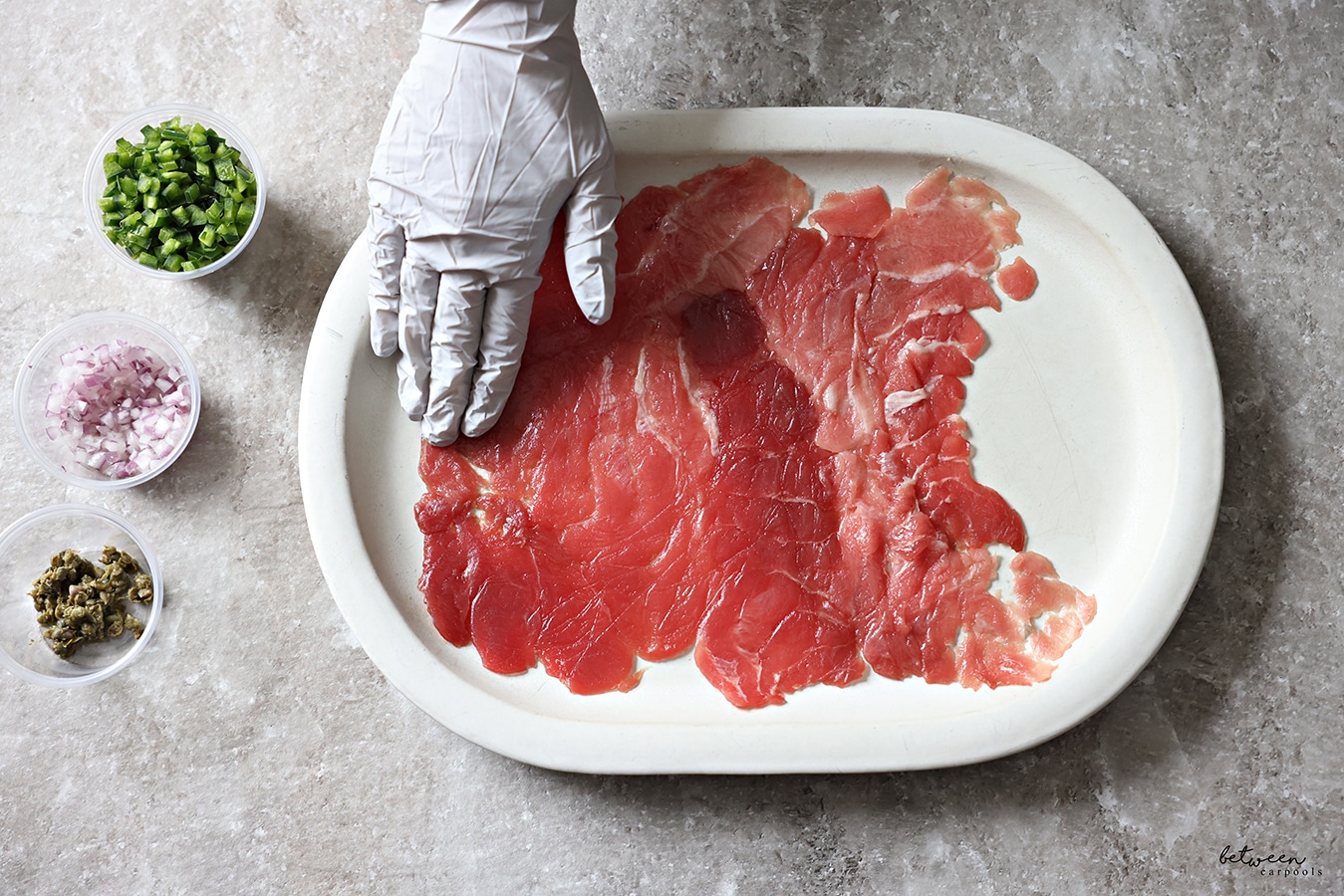 Using a gloved hand to make sure the entire surface of the serving plate is spread evenly with the tuna carpaccio. Diced jalapeño, red onion, and capers, in condiment cups.