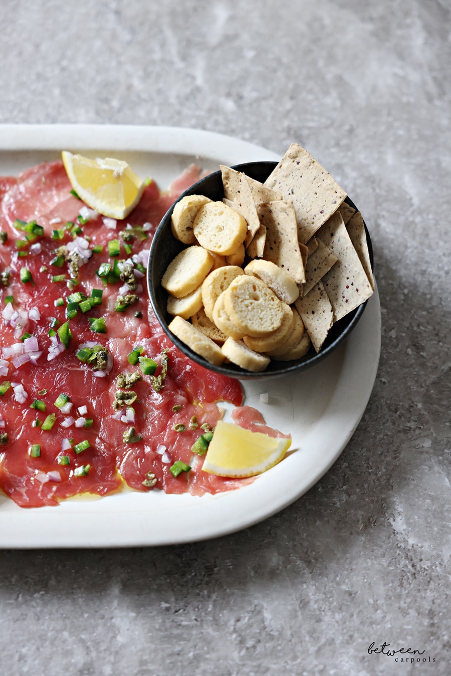 Closeup of tuna carpaccio. with lemon wedges and a bowl of crackers.