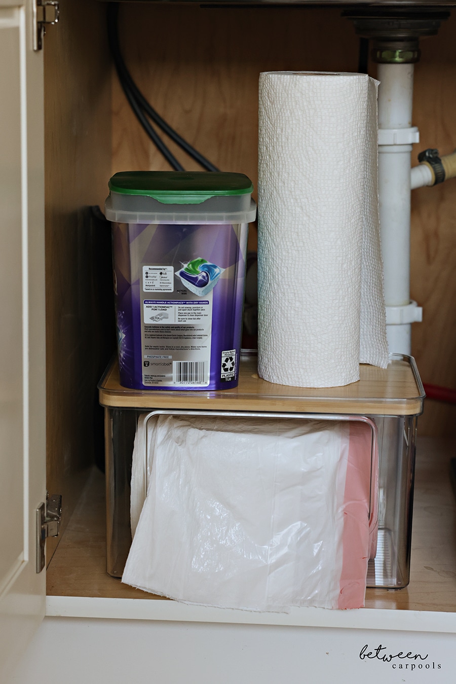 Stackable garbage bag dispenser in the cabinet under the kitchen sink with a roll of paper towel and dishwasher pods on the lid.
