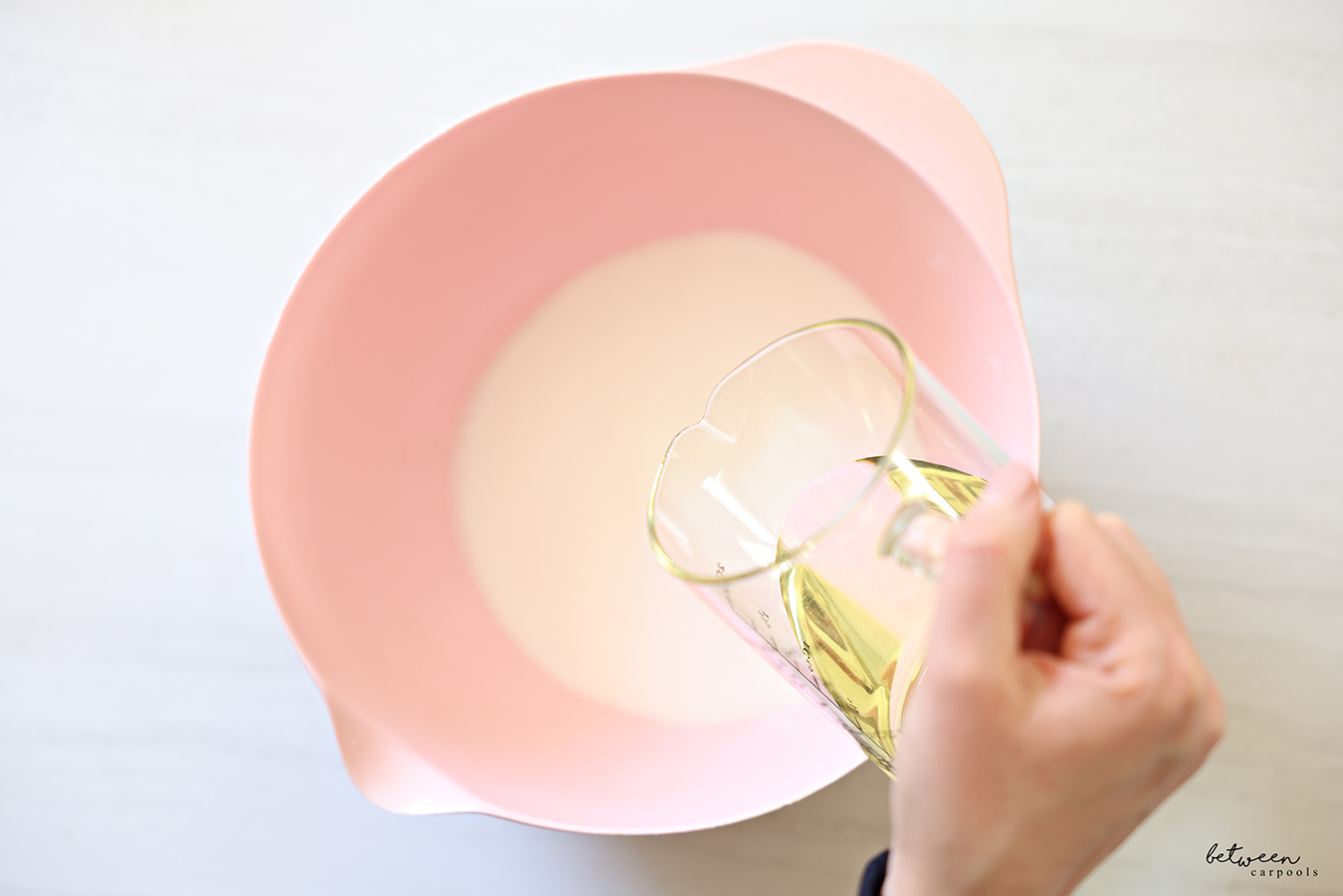 Pouring oil into a pink mixing bowl with spout with sugar.