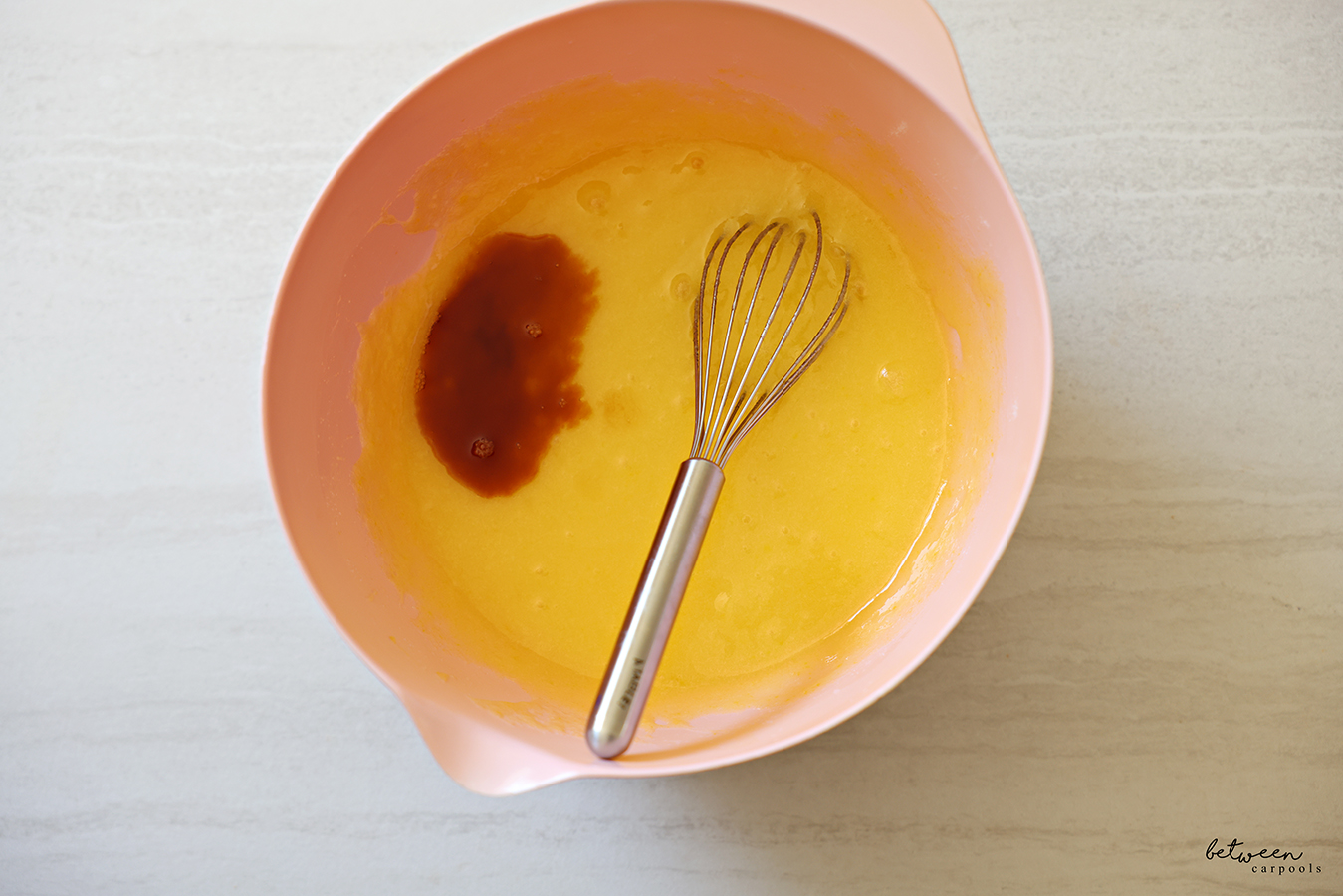 Vanilla extract added to sugar, oil, and eggs, whisked together in a pink mixing bowl with spout with a whisk.