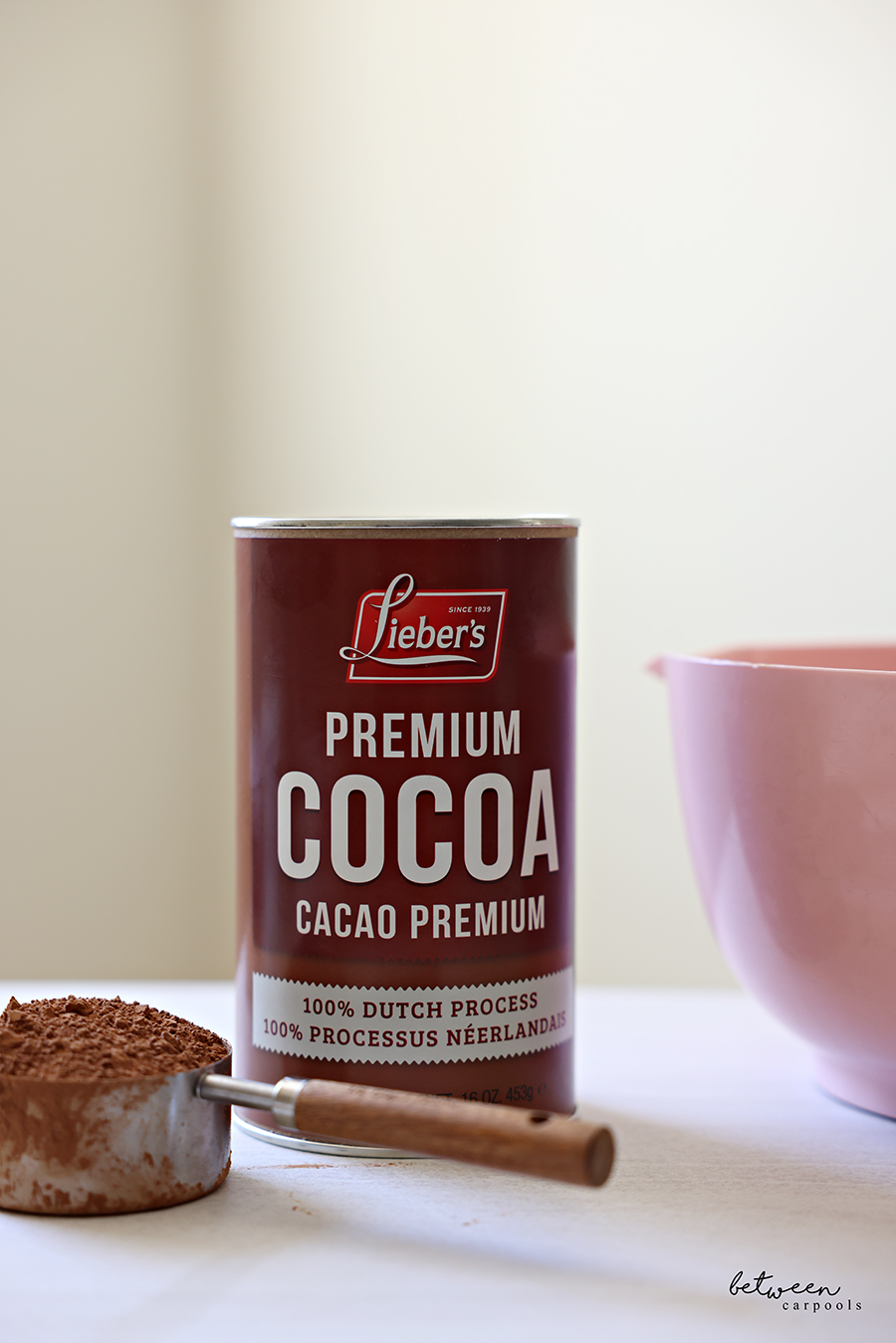 Can of Lieber's Premium Cocoa. A measuring spoon with cocoa. Pink mixing bowl with spout in background.