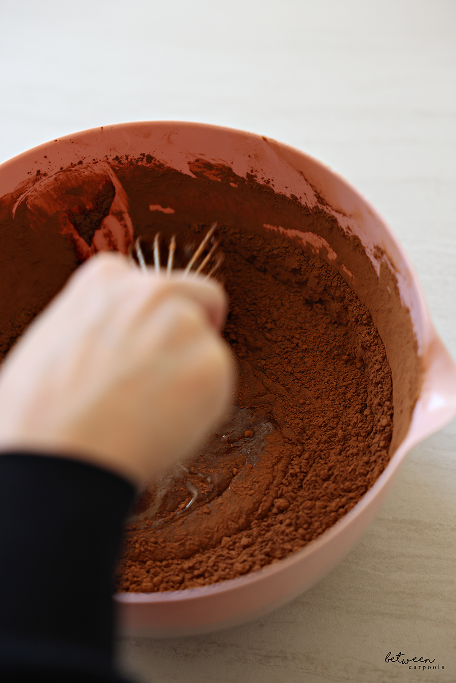 Whisking cocoa into sugar, oil, eggs and vanilla, whisked together in a pink mixing bowl with spout with a whisk.