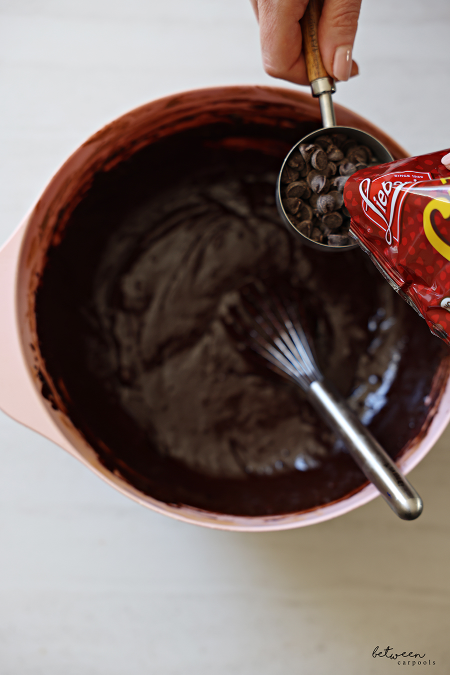 Adding ½ cup Lieber’s chocolate chips to chocolate batter in a pink mixing bowl with spout with a whisk.