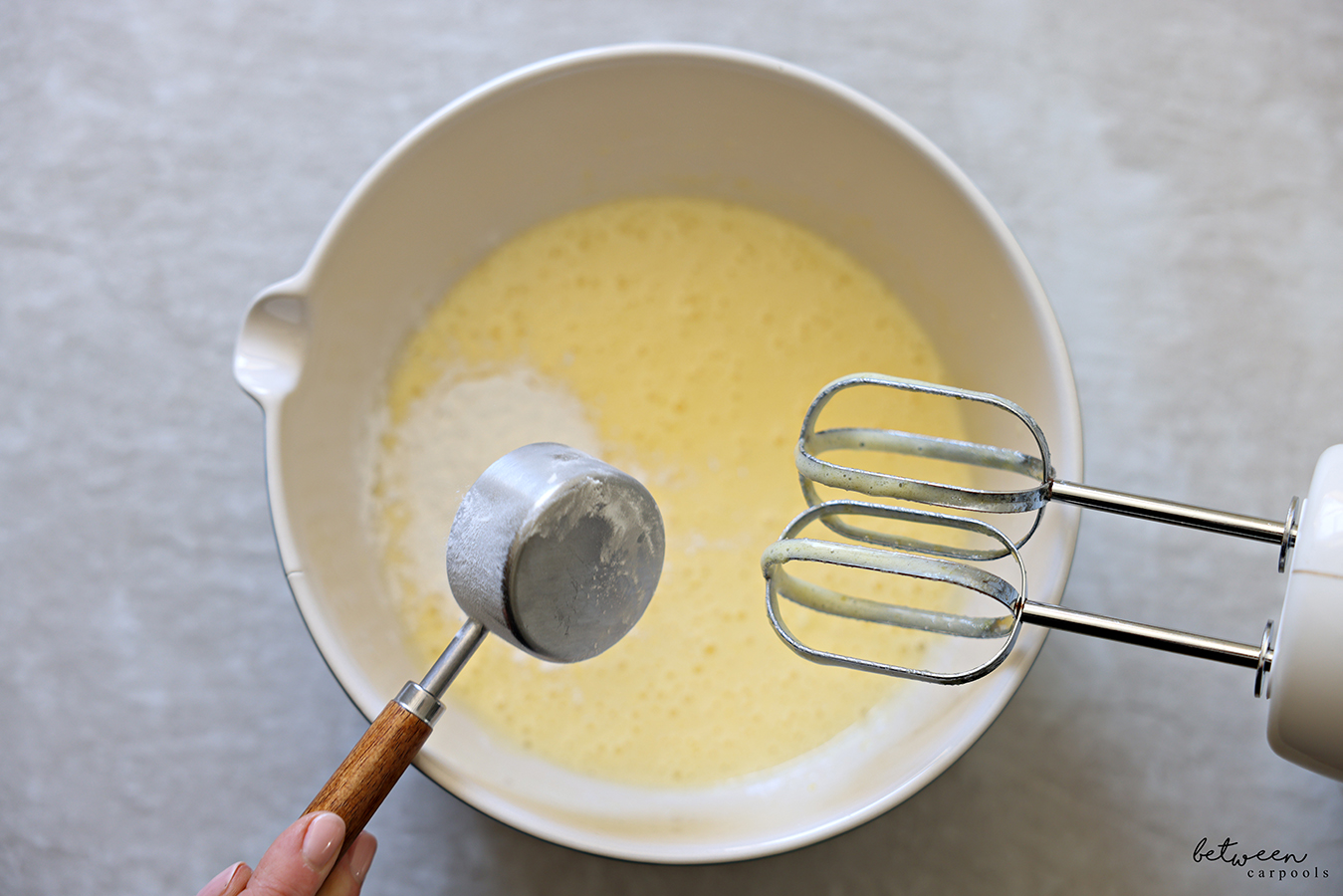Adding potato starch to a creamy mixture of eggs and sugar in a mixing bowl with spout. Hand Mixer beaters leaning on the bowl rim.