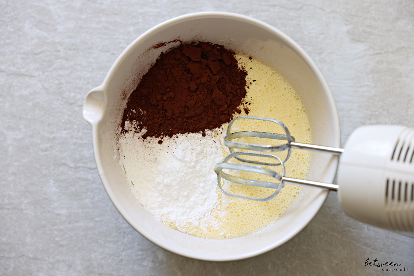 Potato starch and cocoa added to a creamy mixture of eggs and sugar in a mixing bowl with spout. Hand Mixer leaning on the bowl rim.