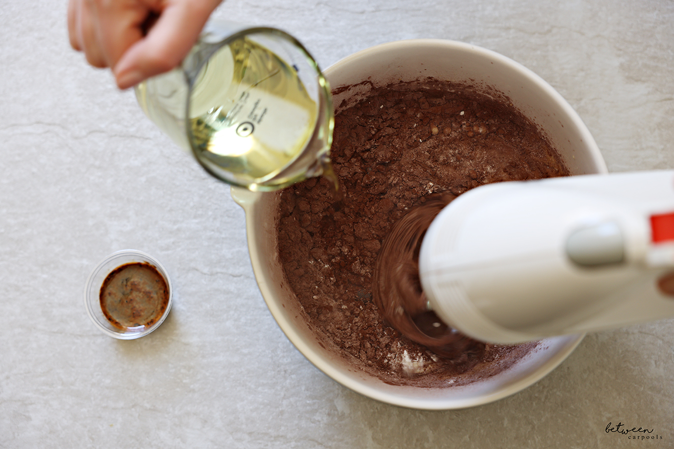 Drizzling oil into chocolate batter being beaten with a hand mixer.