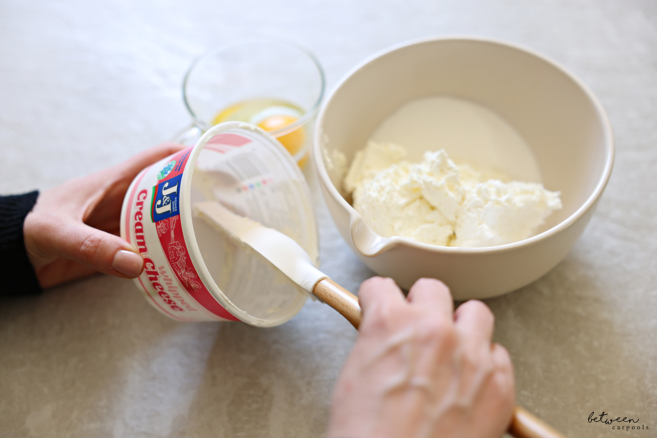 Emptying a J&J whipped cream cheese container with a spatula. Cream cheese and sugar in a mixing bowl with spout. Egg in a glass nearby.